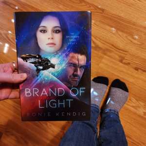 blogger is holding a hardcover copy of Brand of Light with a floor and two socked feet pictured in the background.